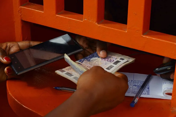 Mains tenant un téléphone et des billets de francs CFA au-dessus d'un carnet, sur un comptoir orange